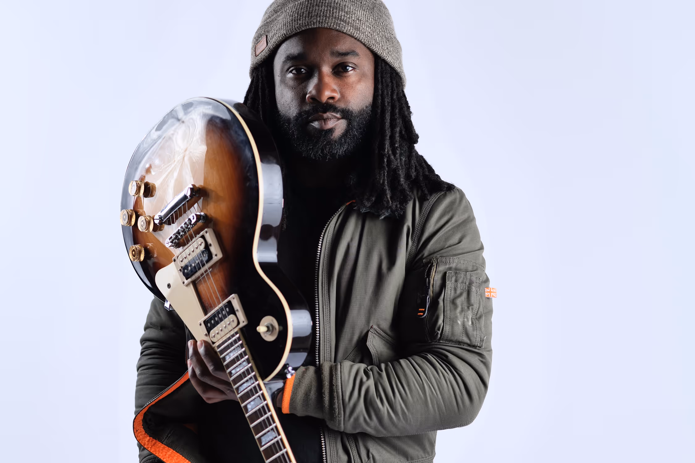 Man with dreadlocks wearing a knit beanie and green jacket holding an electric guitar against a plain light background.