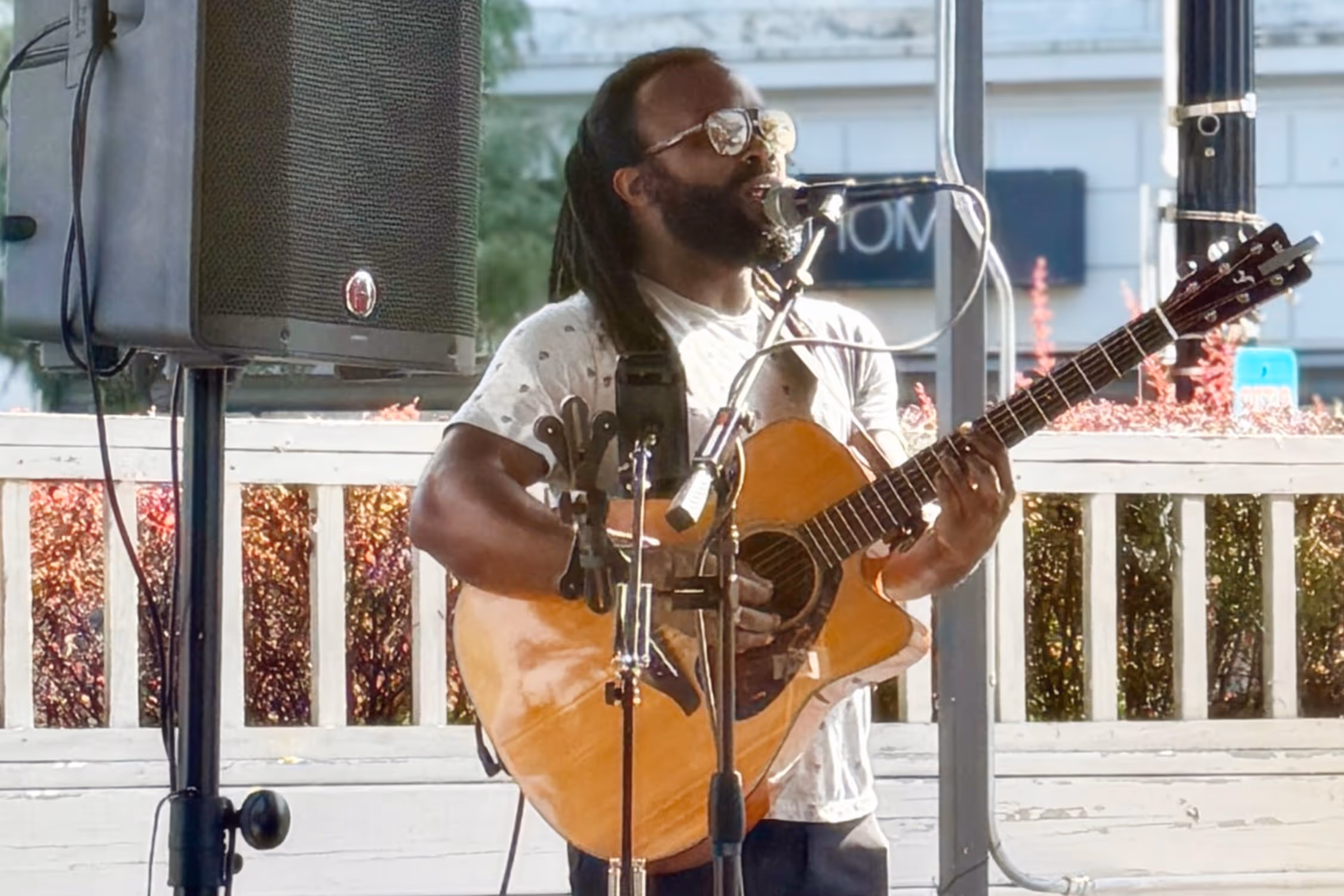 Bearded man with dreadlocks playing acoustic guitar and singing into microphone outdoors.