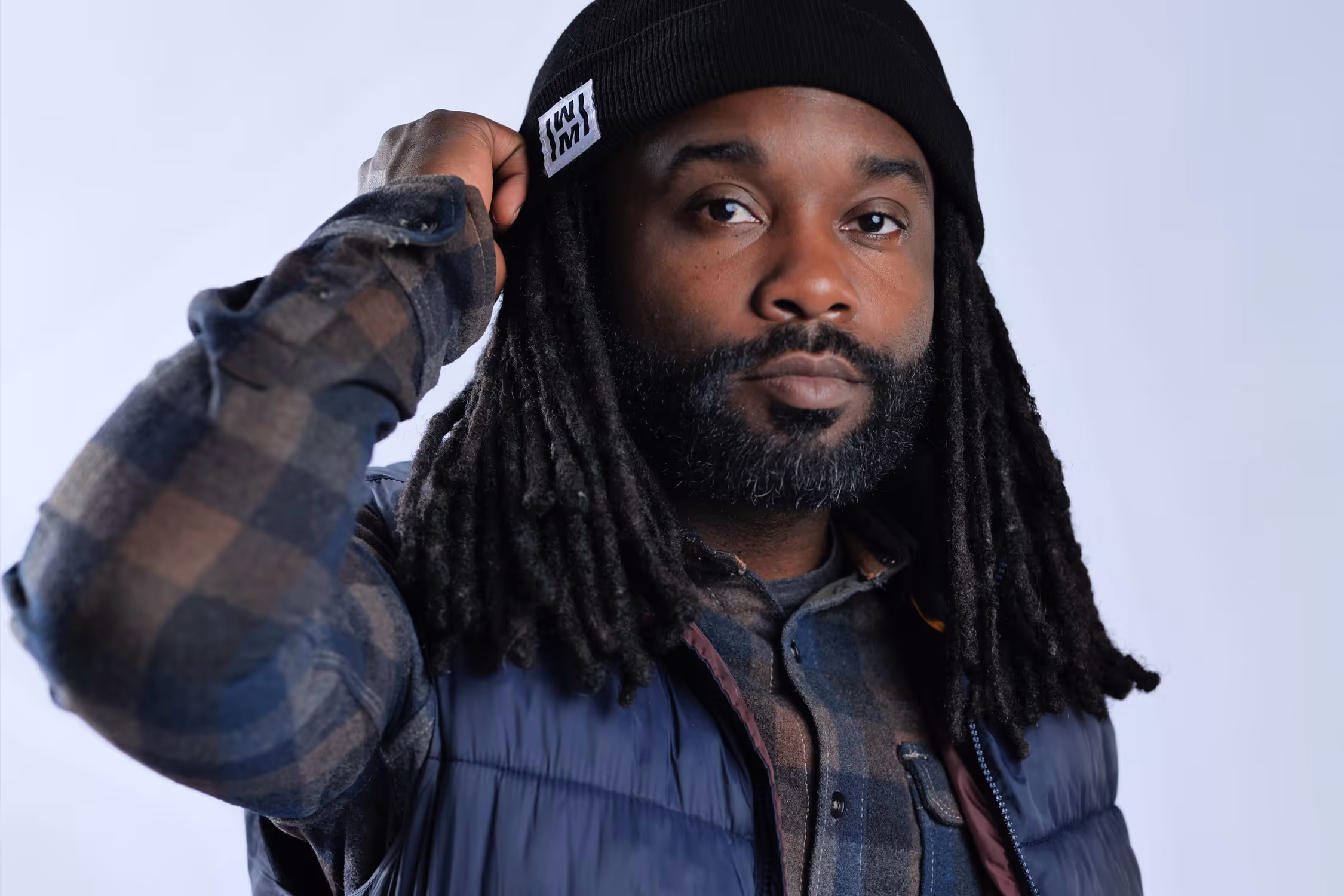 Man with dreadlocks wearing a black beanie, blue puffer vest, and plaid shirt adjusting his hat.