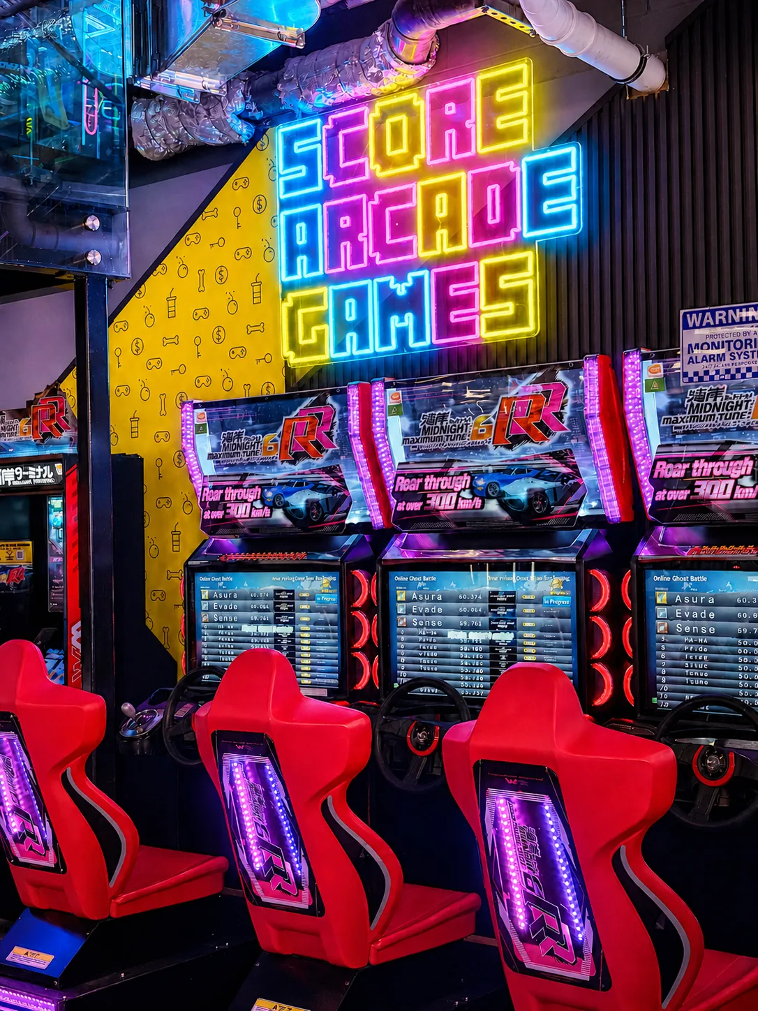 Row of racing arcade machines beneath a neon Score Arcade Games sign at KOKO Amusement
