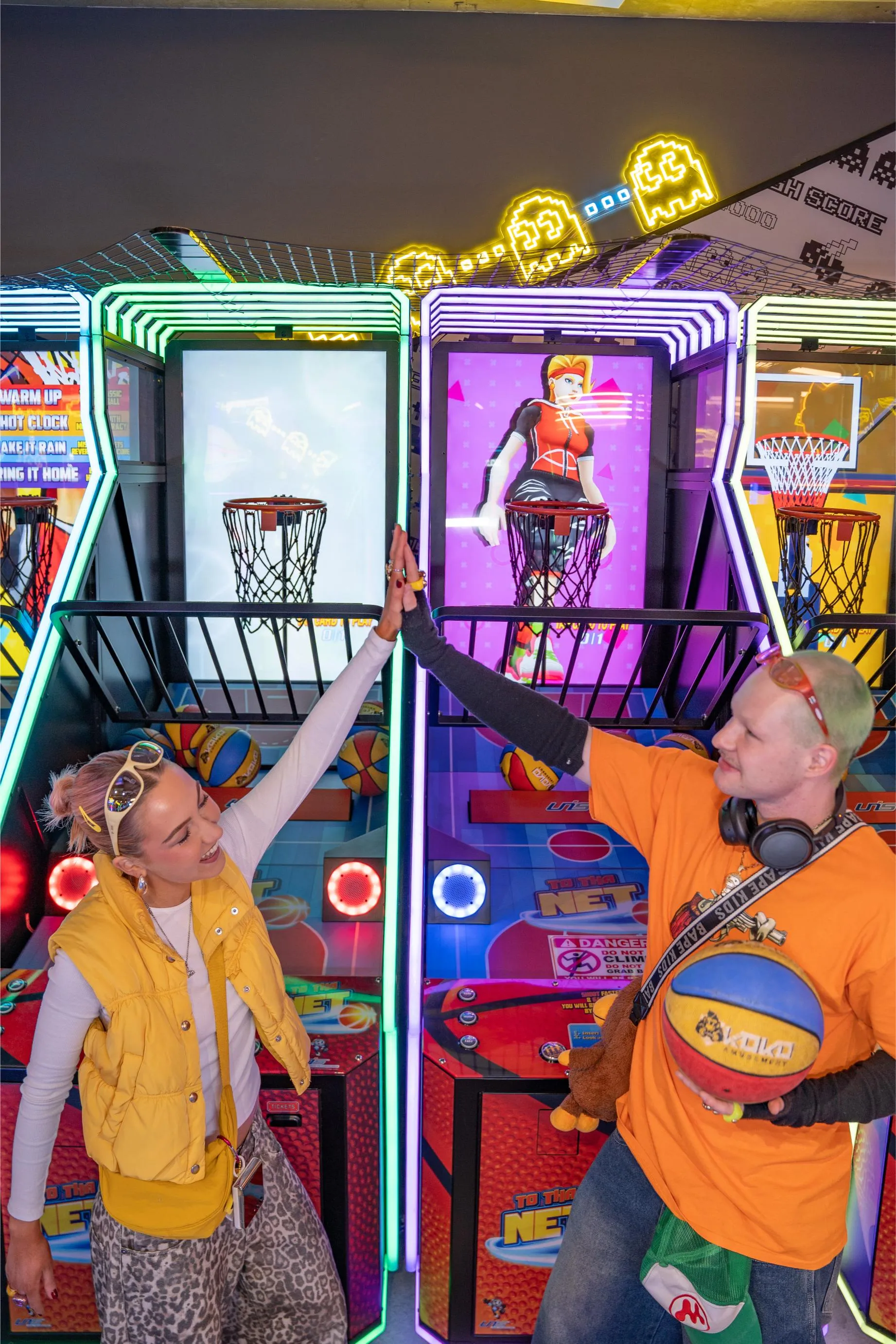 Two friends high-fiving in front of neon basketball arcade machines at KOKO Amusement