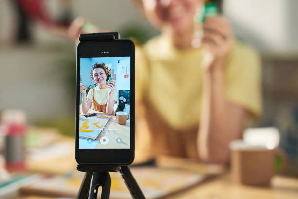 female influencer recording a video of her showing paintbrush and gouache Focus on smartphone screen with happy young female artist showing paintbrush and small jar of gouache during online masterclass