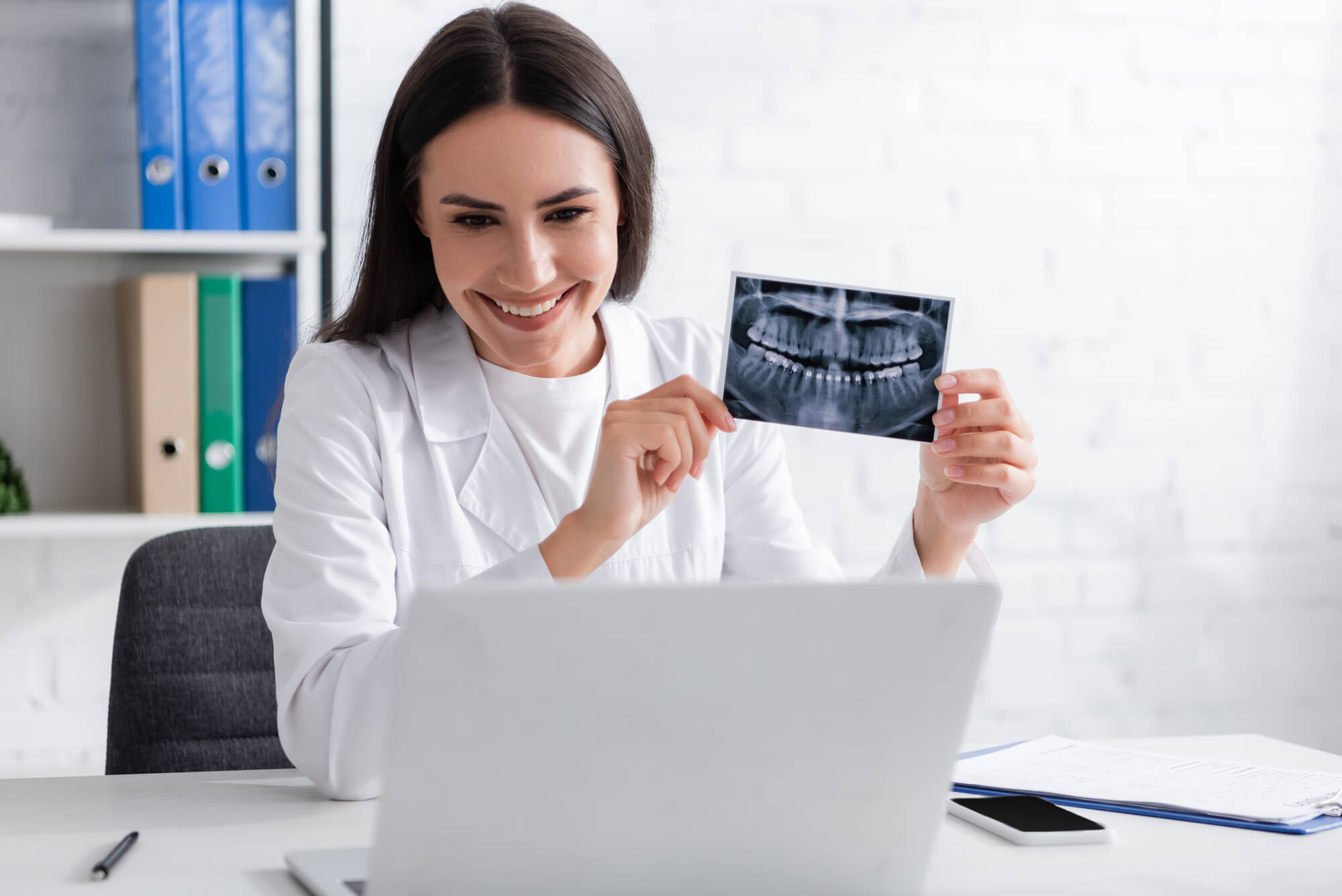 smiling dentist holds up a dental X-ray while conducting a virtual consultation using her laptop