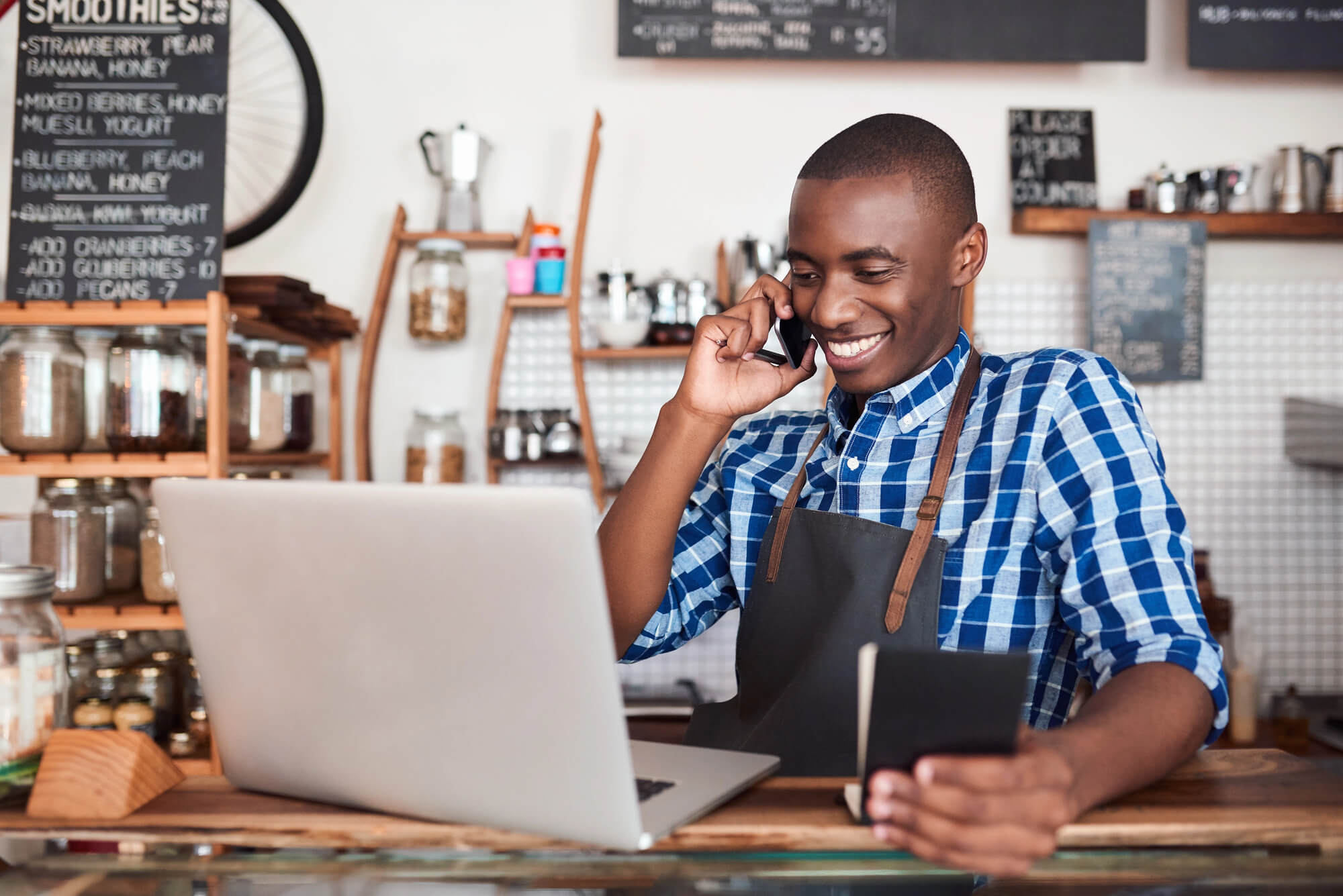 a smiling cafe owner in an apron using a laptop while talking on the phone 