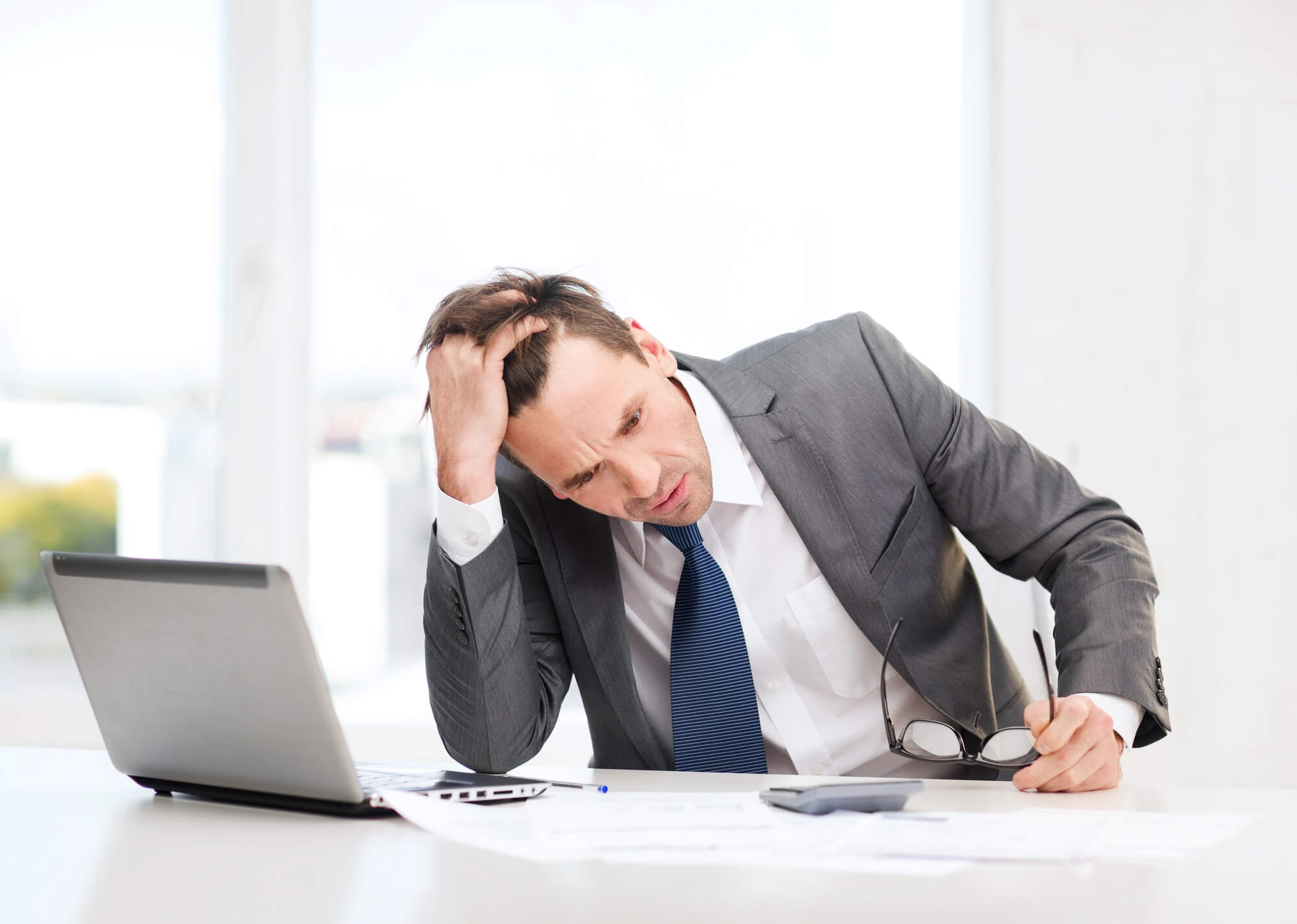 a stressed business leader sitting at his desk, holding his head while looking at documents and a laptop.