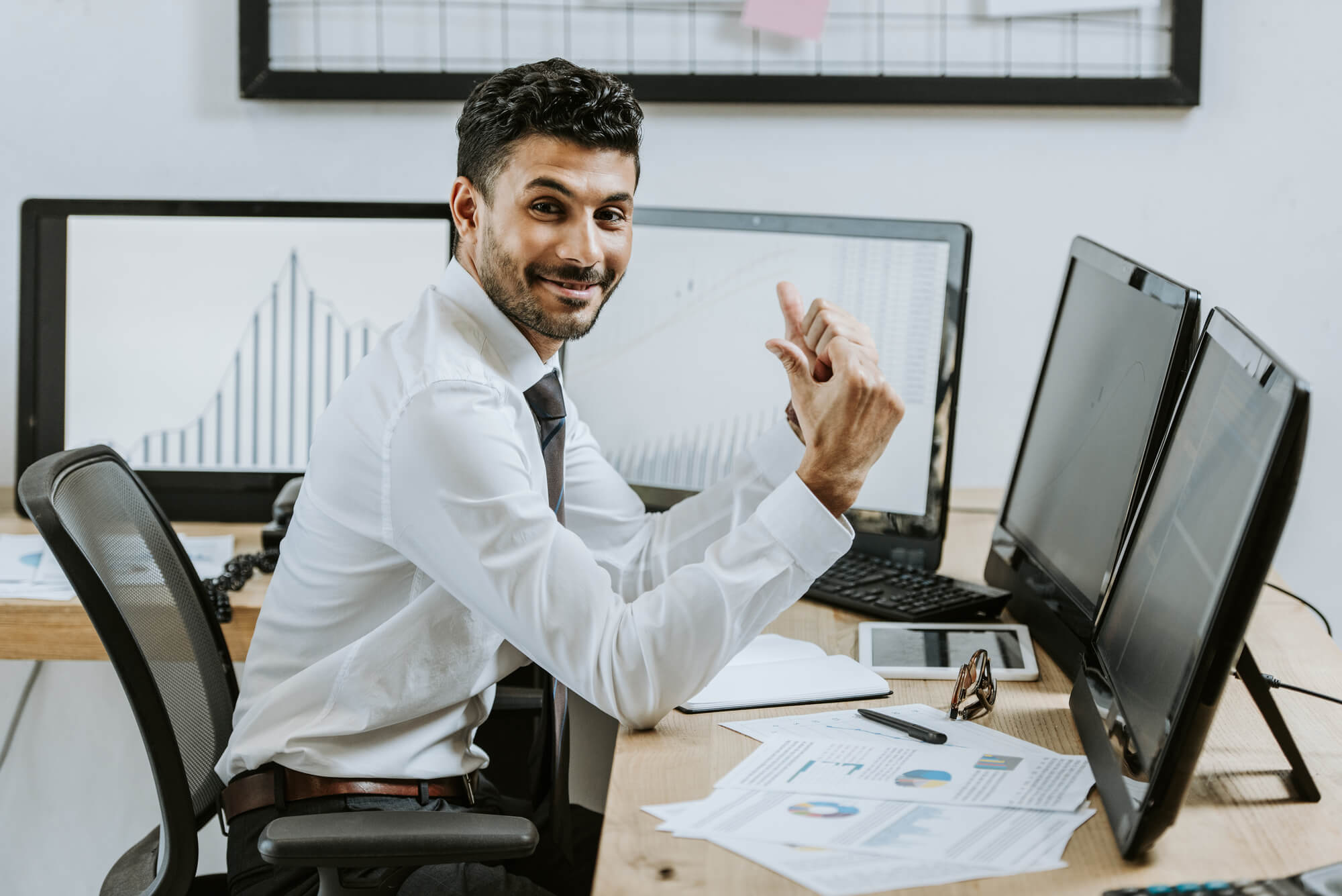a smiling businessman at a desk with multiple monitors giving a thumbs up
