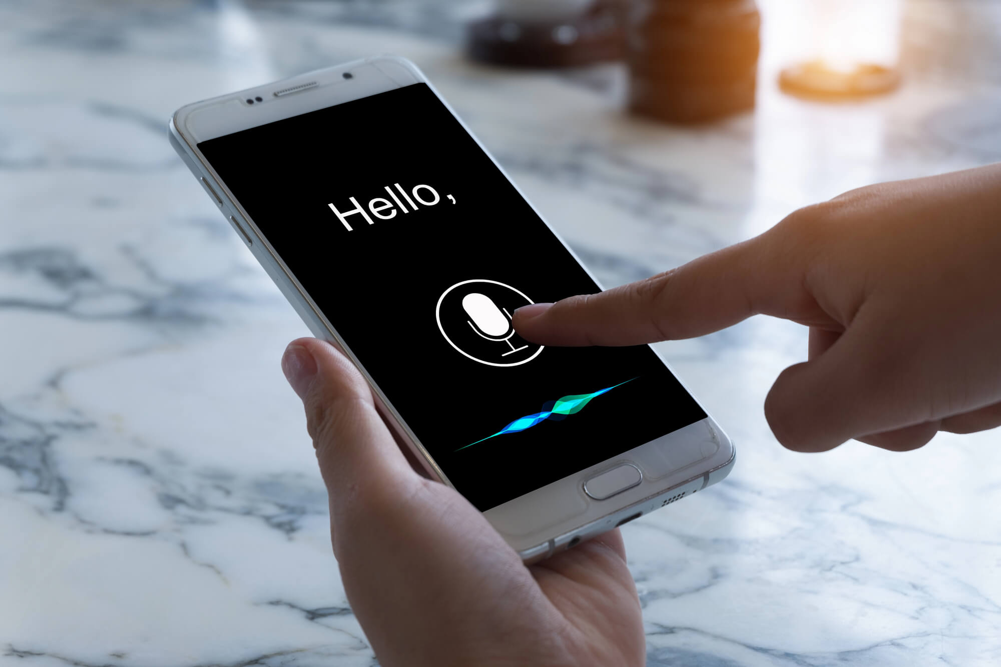 a hand holding a smartphone while tapping the voice assistant icon on a screen that says "Hello."