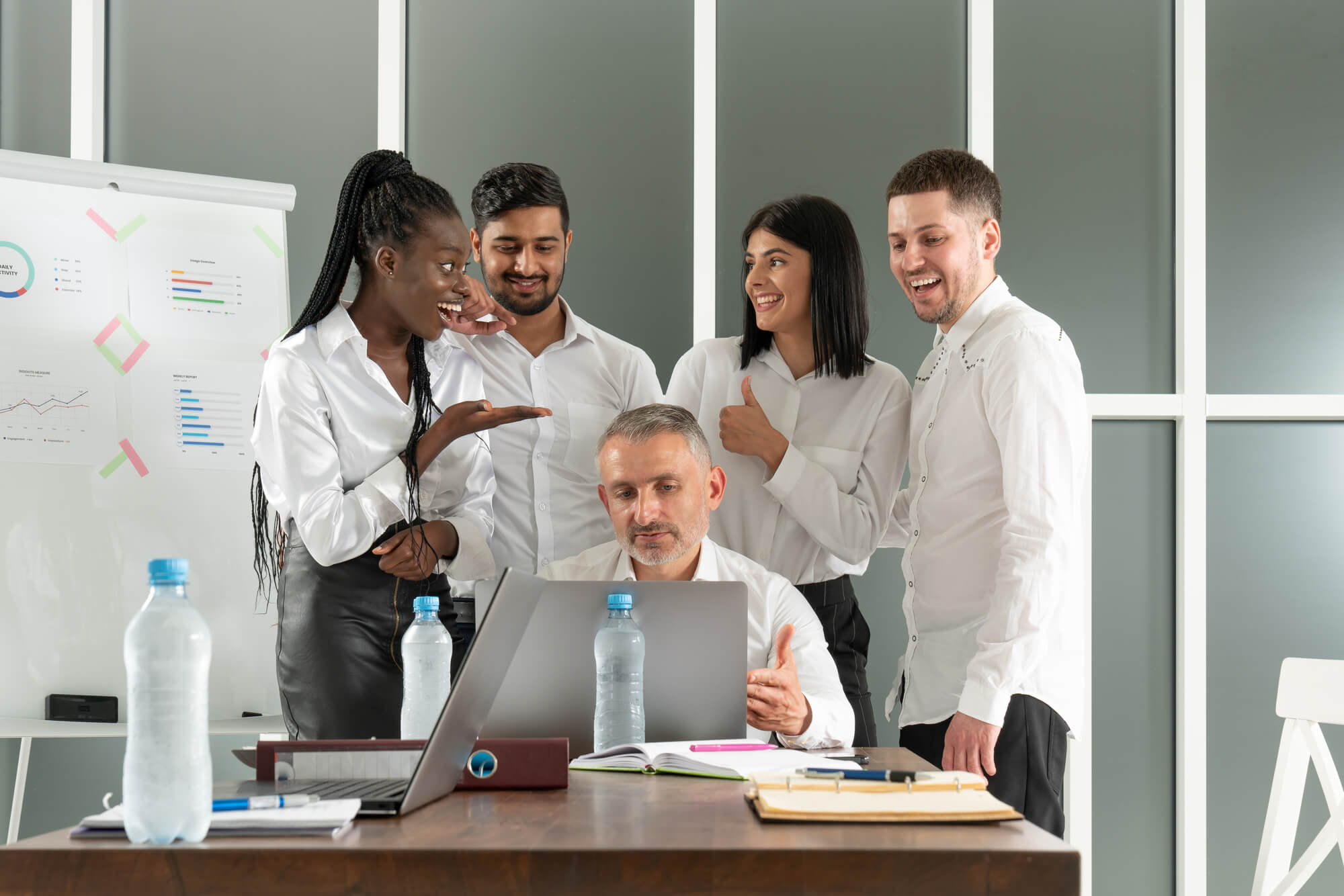 a manager works on a laptop while his team of young employees gathers around, engaged in discussion