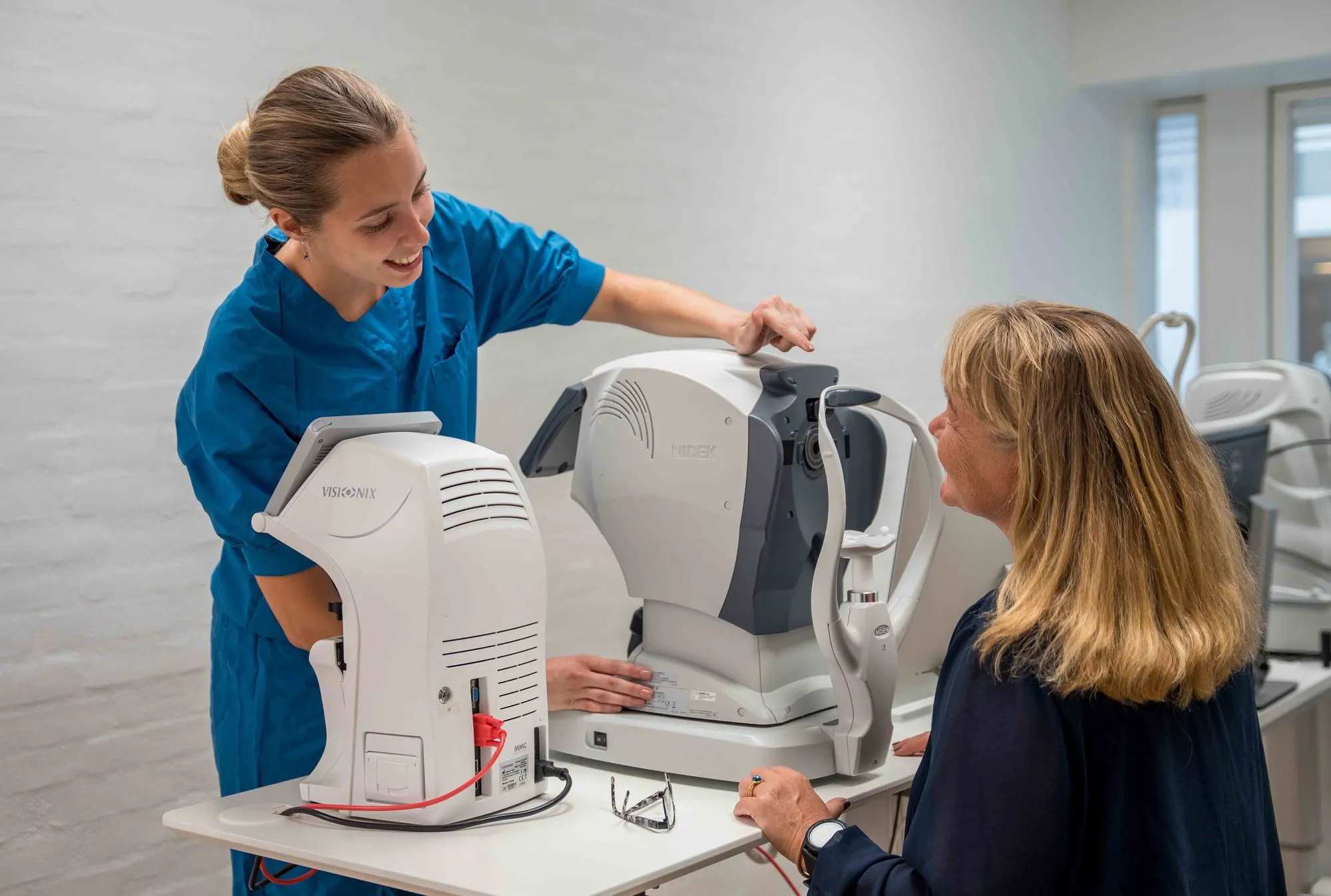 En sundhedsperson i blå uniform hjælper en kvinde med en øjenundersøgelsesmaskine i et klinikrum.