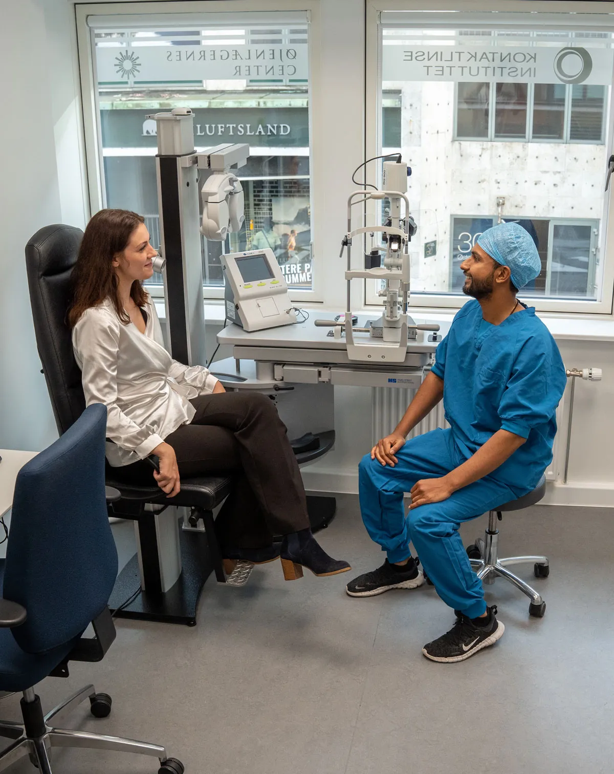 Kvinde i hvid skjorte sidder på en stol i en øjenklinik og taler med en mandlig øjenlæge i blå scrub og kirurgisk hætte.