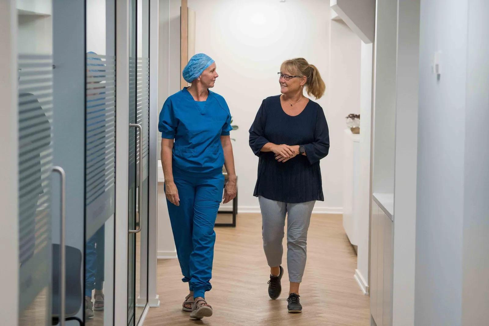 To kvinder, en i blå hospitalsuniform og en i casual tøj, går og taler i en lys hospitalsgang.