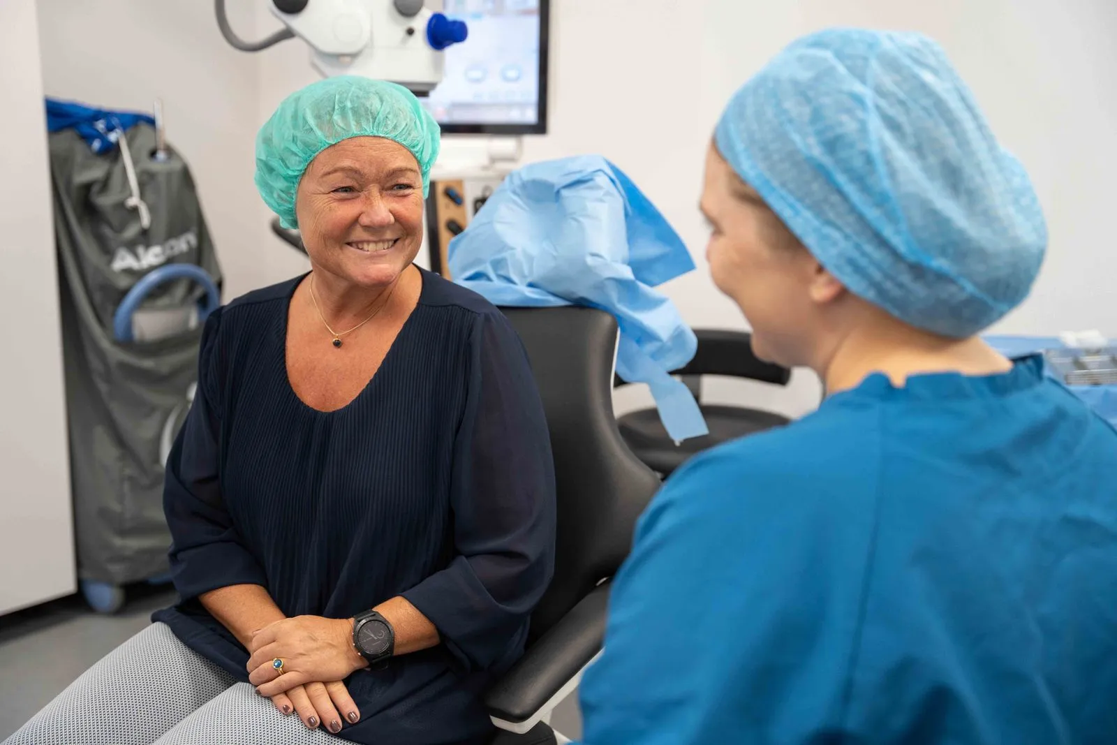 Kvinde med grøn operationshætte smiler til en sundhedsprofessionel i blå uniform med blå operationshætte i et klinikrum.