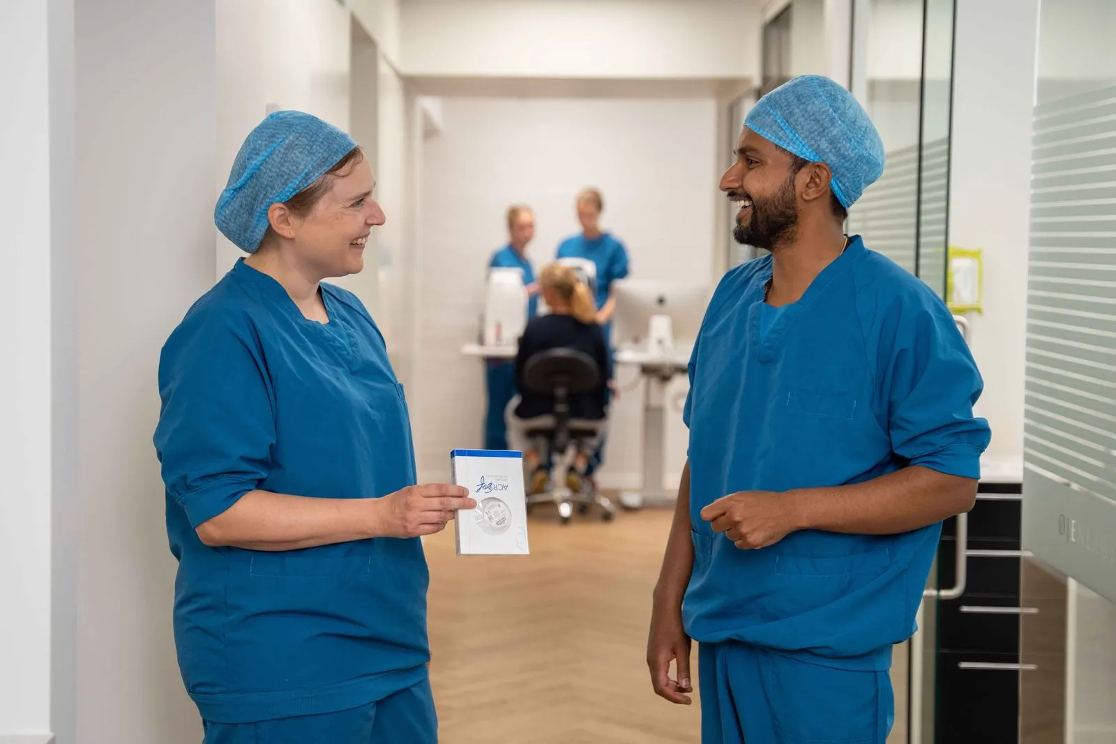 To sundhedspersonale i blå operationskitler og hårnet smiler og taler sammen i en hospitalsgang.