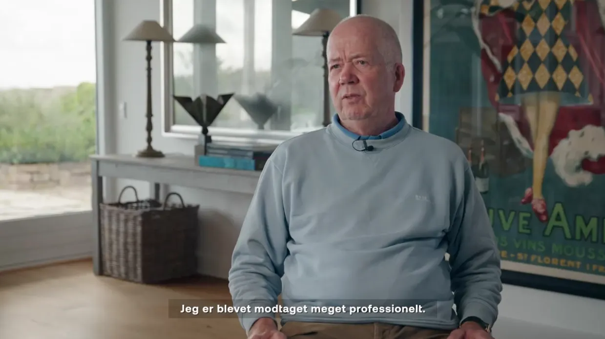 Ældre mand i lyseblå sweatshirt sidder indendørs foran et bord med lamper og en kurv, med en stor farverig plakat i baggrunden.