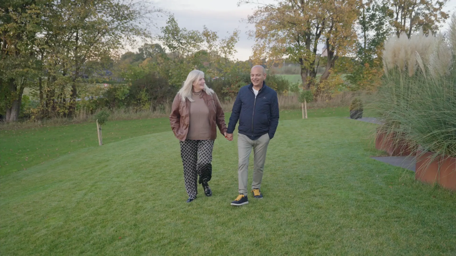 Et par holder hinandens hænder og går på en græsplæne med træer i baggrunden.