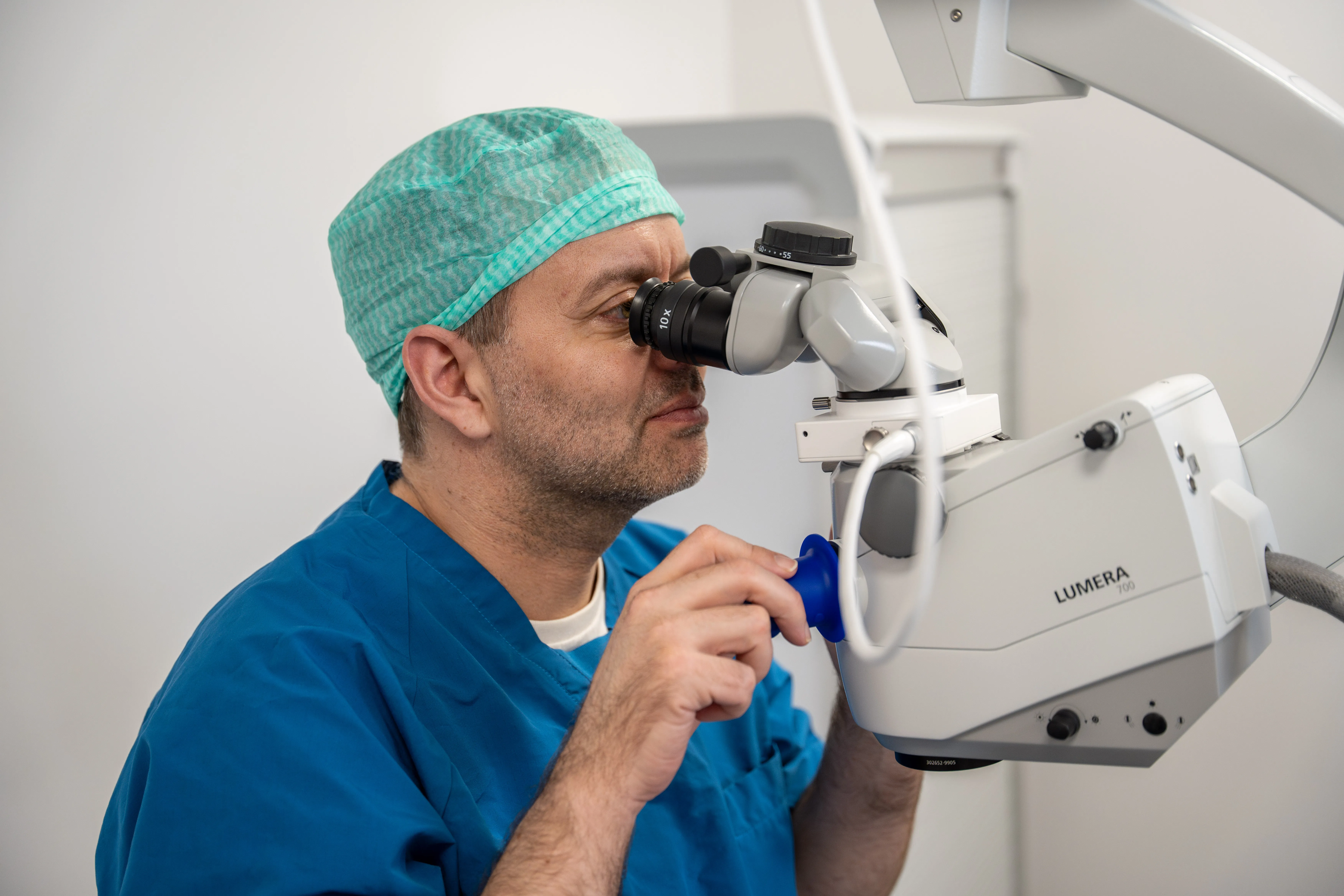 Læge iført kirurgisk hue og blå kitler kigger gennem et mikroskop i et klinisk miljø.