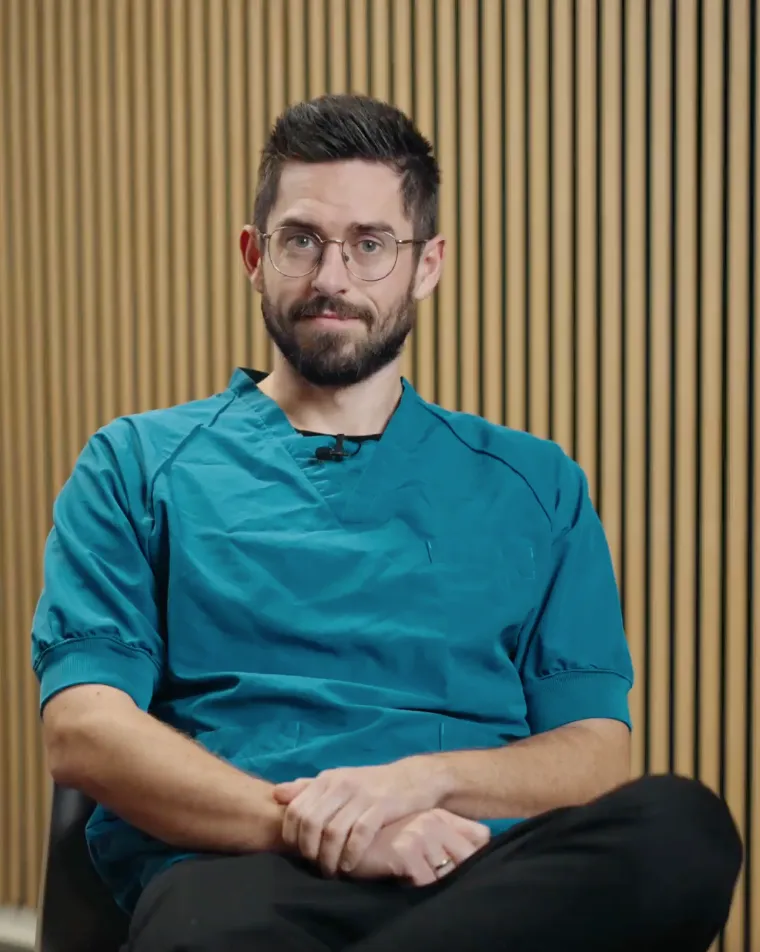 Mand med briller og skæg iført blå medicinsk scrub sidder foran en væg med lyst træpanel.