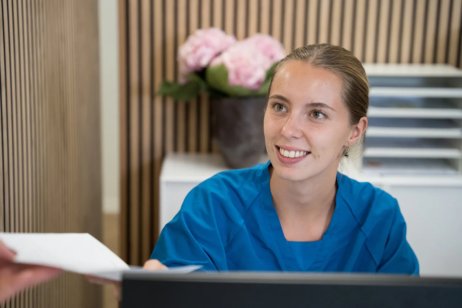 Sundhedspersonale iført blå scrubs smilende og tager imod et dokument ved en receptionistdisk.