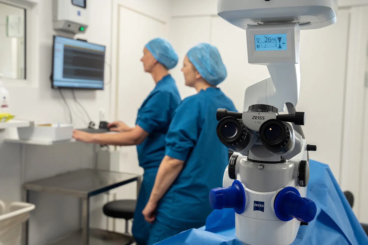 To sundhedspersonale i blå operationsdragter og hårnet arbejder ved en computerskærm i et klinikværelse med et kirurgisk mikroskop i forgrunden.