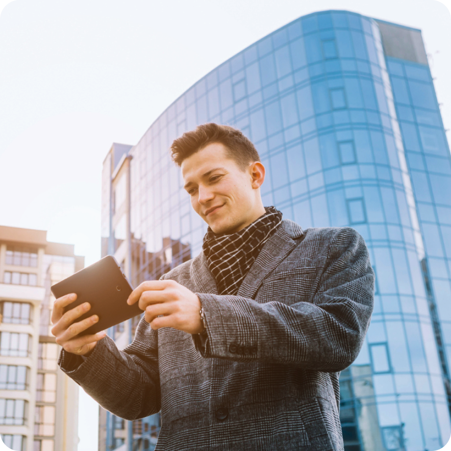 Man in a plaid coat and scarf using a tablet outside with modern glass buildings in the background.