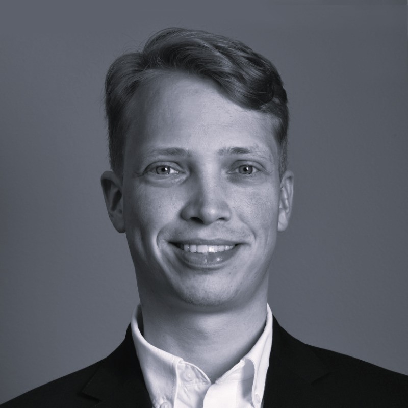 Smiling young man with short hair wearing a white shirt and dark blazer against a plain background.