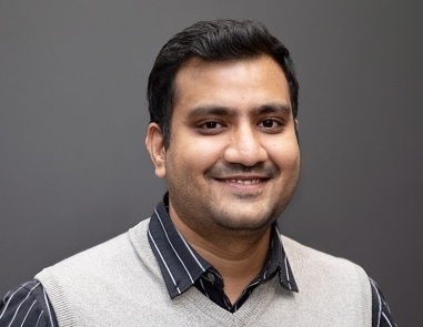 Smiling man with short black hair wearing a striped black shirt and light gray vest against a gray background.