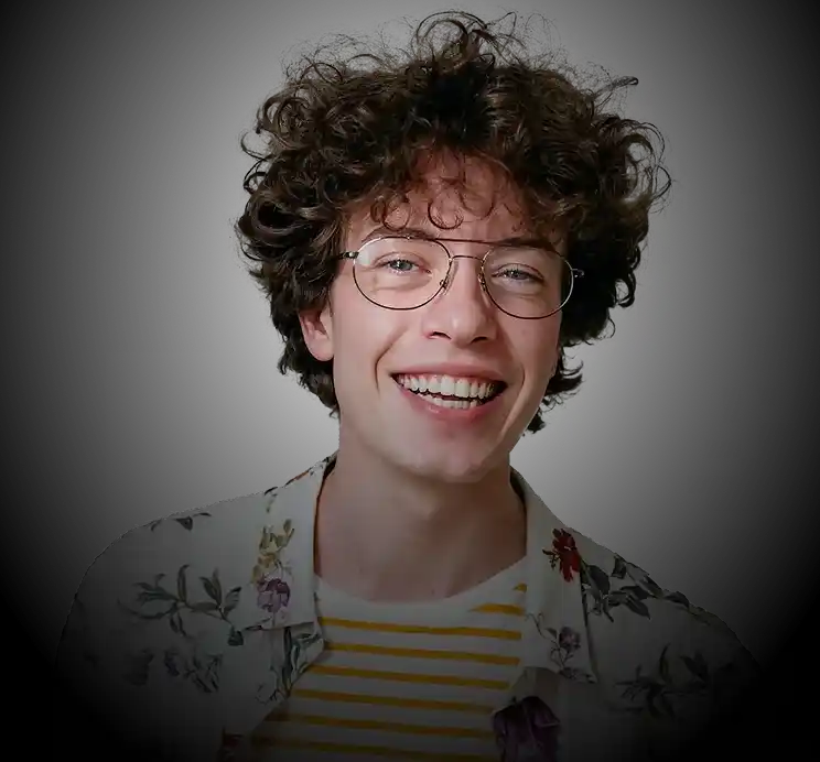 Smiling young man with curly hair and glasses wearing a floral shirt and striped t-shirt against black background.