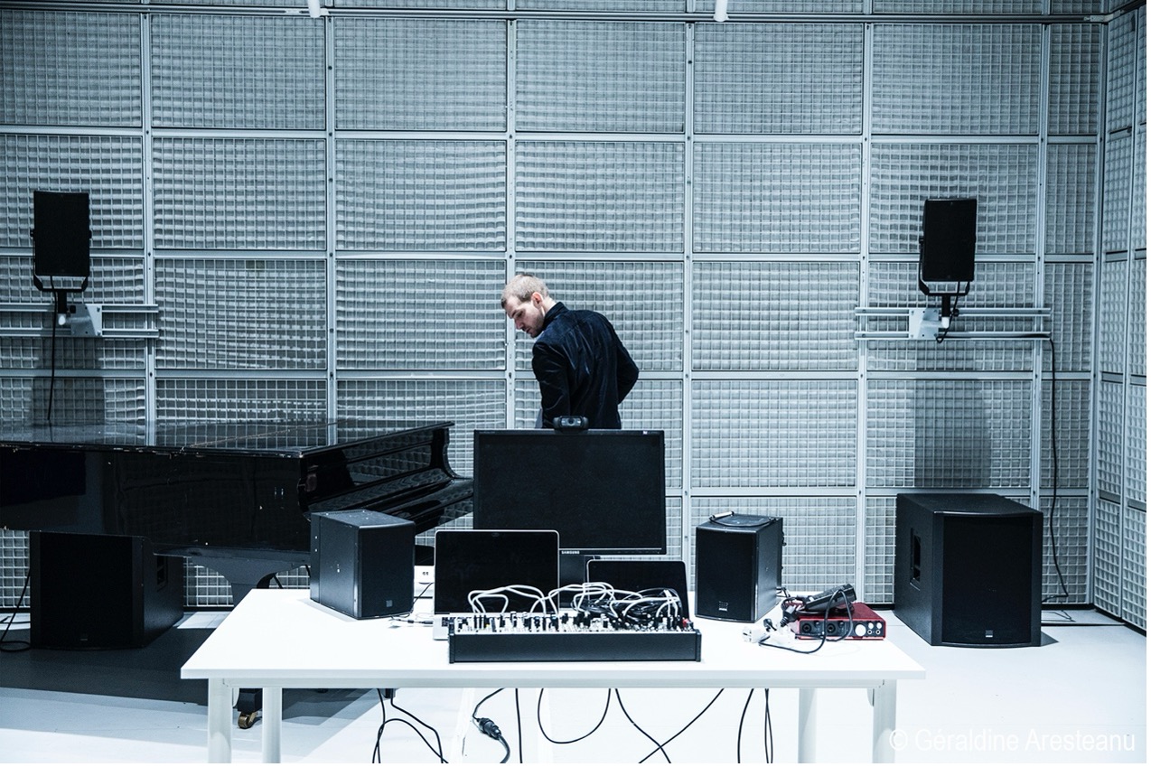 Man in a black jacket standing near a grand piano in a soundproofed room with audio equipment on a white table in the foreground.