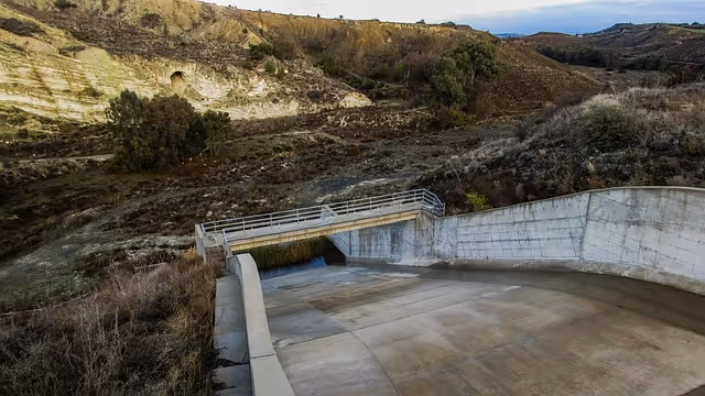 Concrete spillway structure in a dry, hilly landscape with sparse vegetation under a partly cloudy sky.
