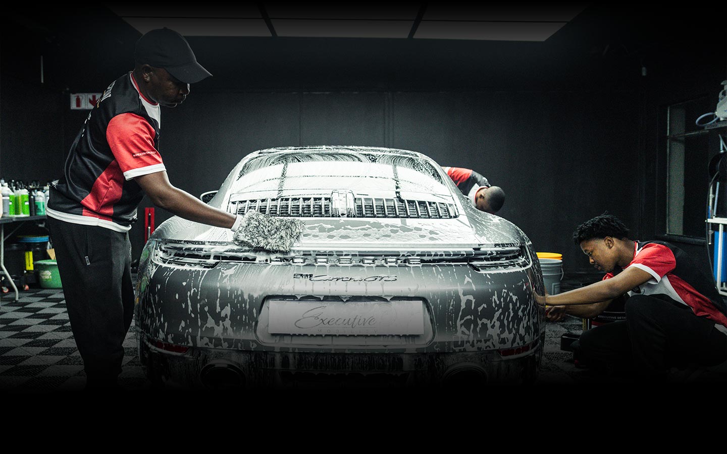 Three workers wearing black and red uniforms washing and detailing a silver Porsche Carrera GTS covered in soap suds in a garage.