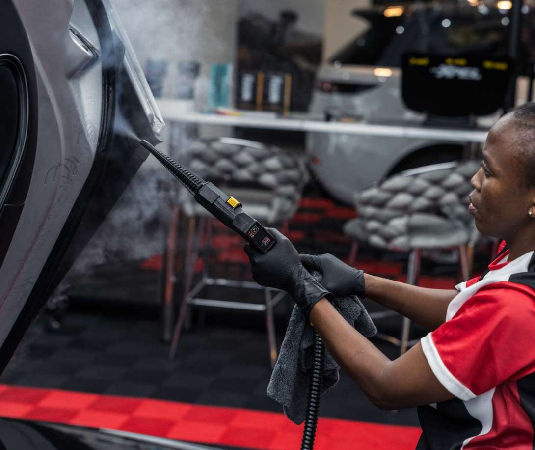 Person wearing black gloves holding a steam cleaner and cleaning the door area of a vehicle indoors.