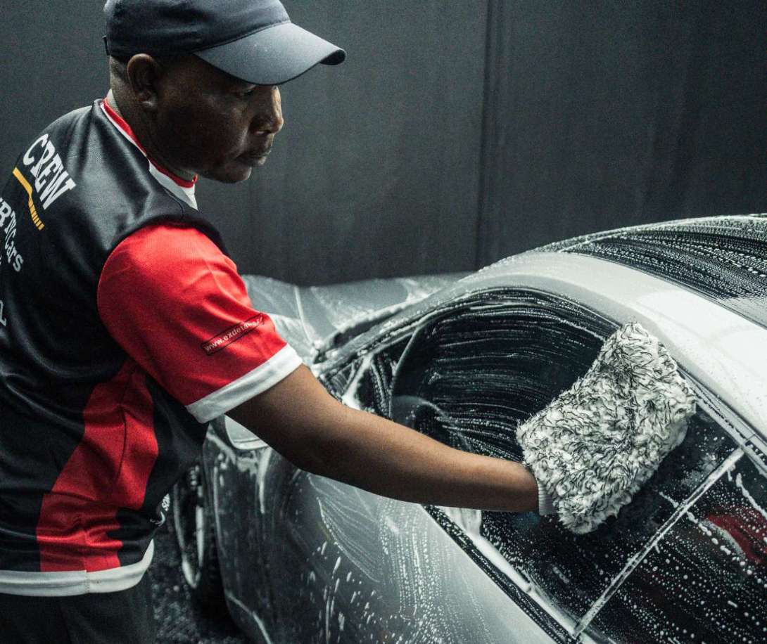 Man wearing a cap and crew vest washing the window of a silver car with a soapy mitt.