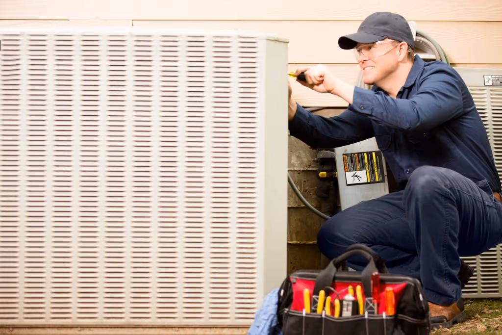 Technician wearing safety glasses and cap fixing an outdoor air conditioning unit with a screwdriver.