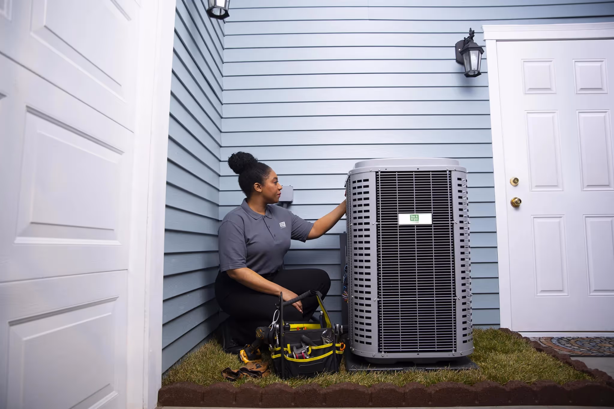 Technician kneeling beside an outdoor air conditioning unit with tools on the grass near a blue house exterior.