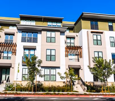Modern three-story residential building with balconies, large windows, and small trees in front under a clear blue sky.