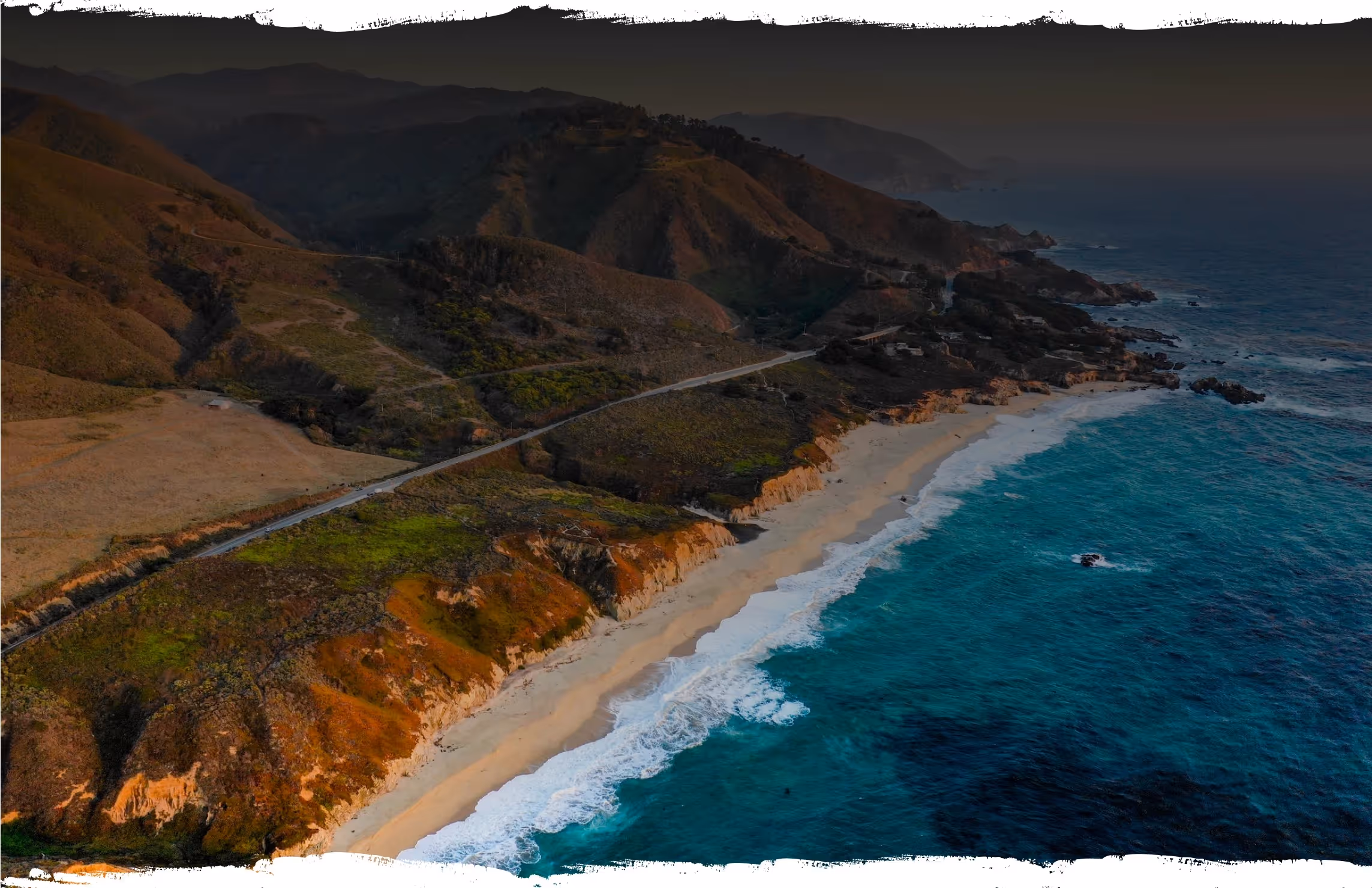 Aerial view of a coastal road winding along rugged cliffs with waves crashing onto a sandy beach and turquoise ocean.