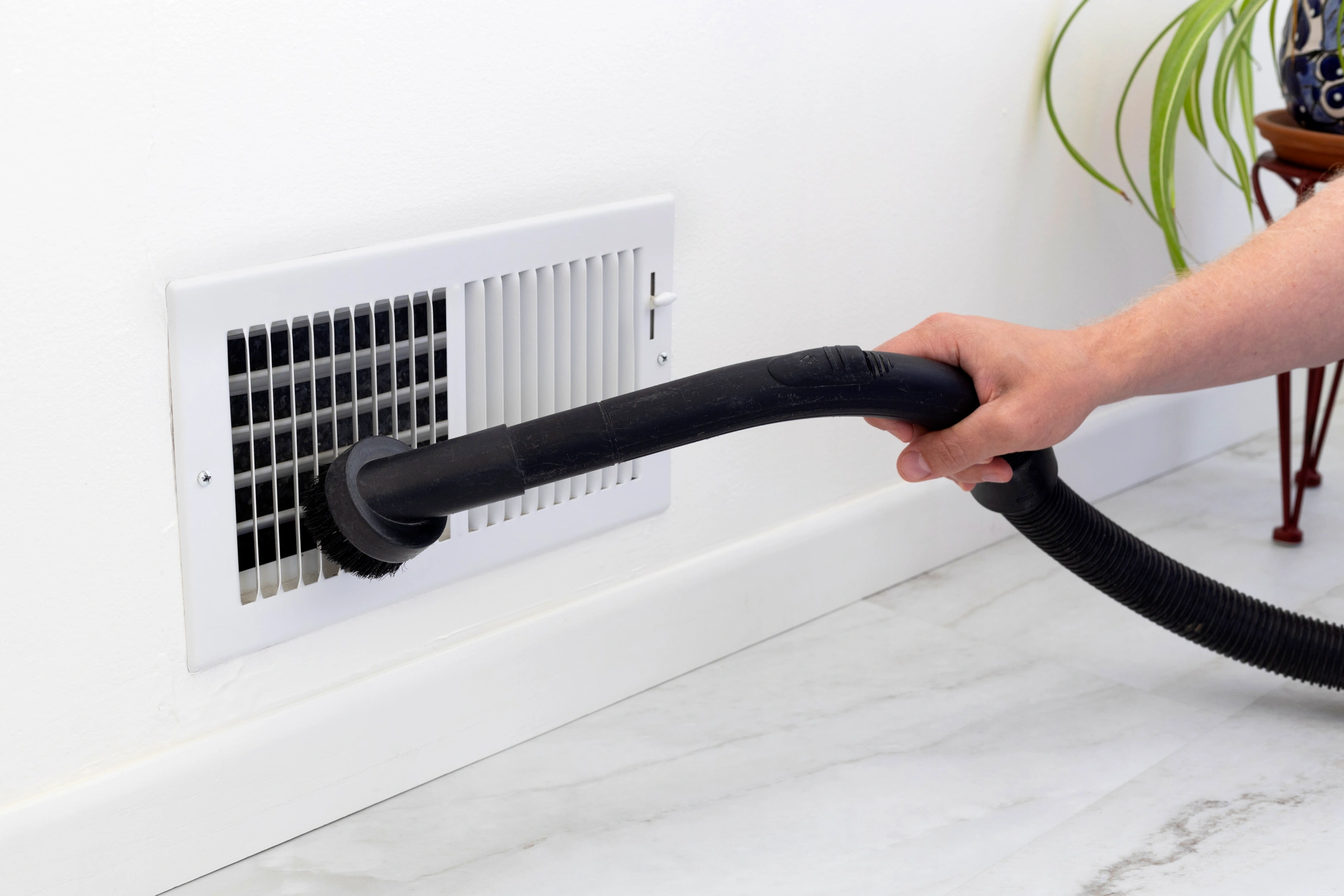 A hand uses a vacuum cleaner attachment to clean a white air vent on a wall. 