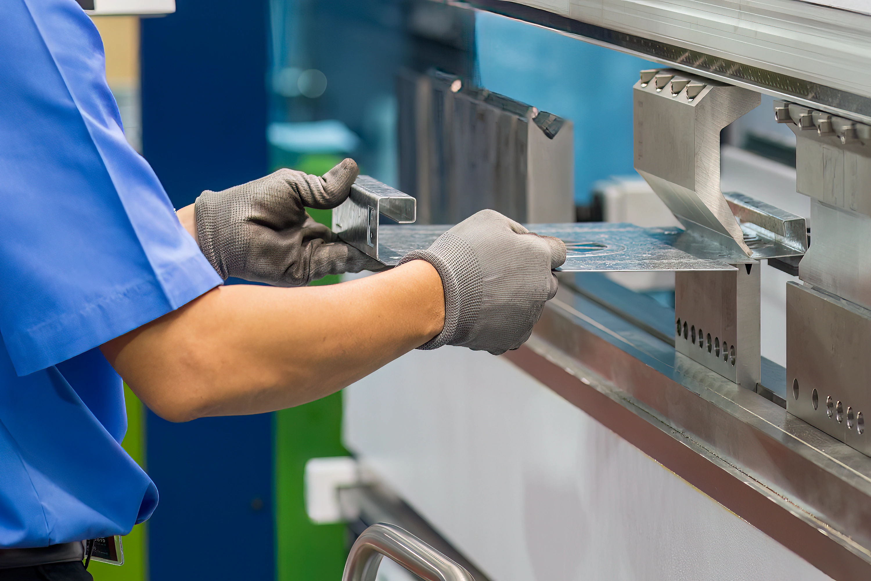 A worker in a blue shirt and gloves carefully bends a metal sheet using an industrial press brake machine