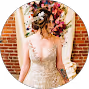 Bride in a lace wedding dress holding a bouquet of flowers, standing against a brick wall.