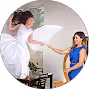 Two women engaging in a playful pillow fight indoors, one wearing a white dress and the other a blue dress.