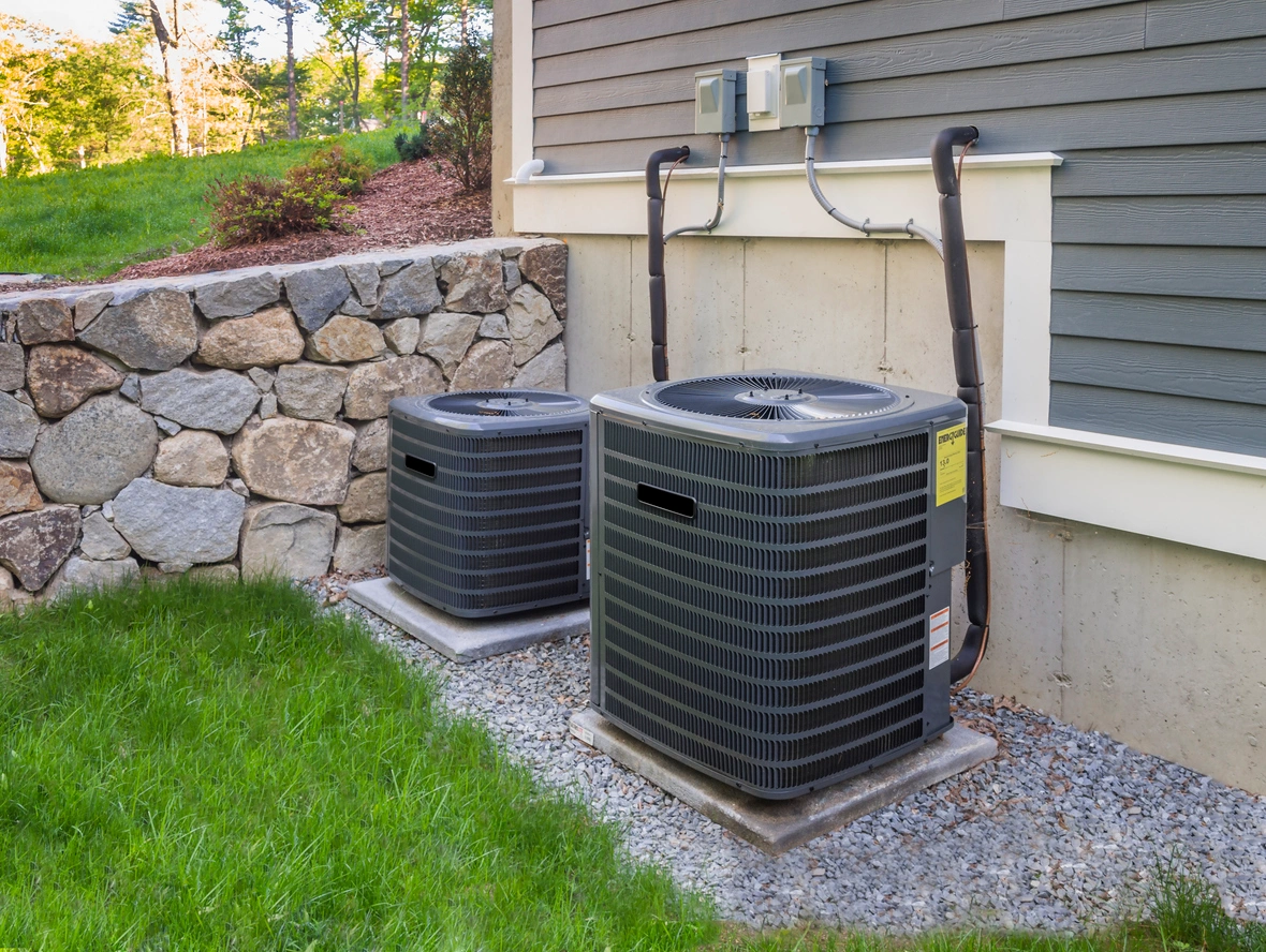 Two outdoor air conditioning units are positioned on concrete slabs next to a house with gray siding. 