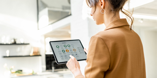 Woman in a brown blazer using a tablet to control smart home devices in a bright kitchen.