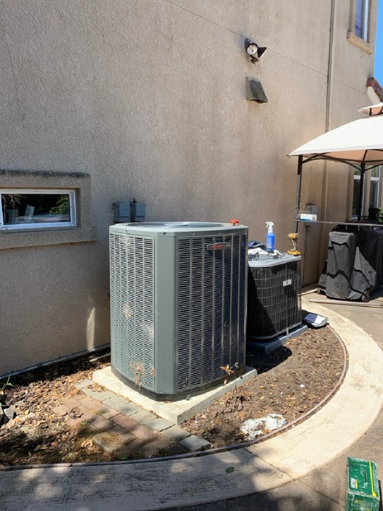 Two outdoor air conditioning units installed next to a beige stucco house wall under sunlight.