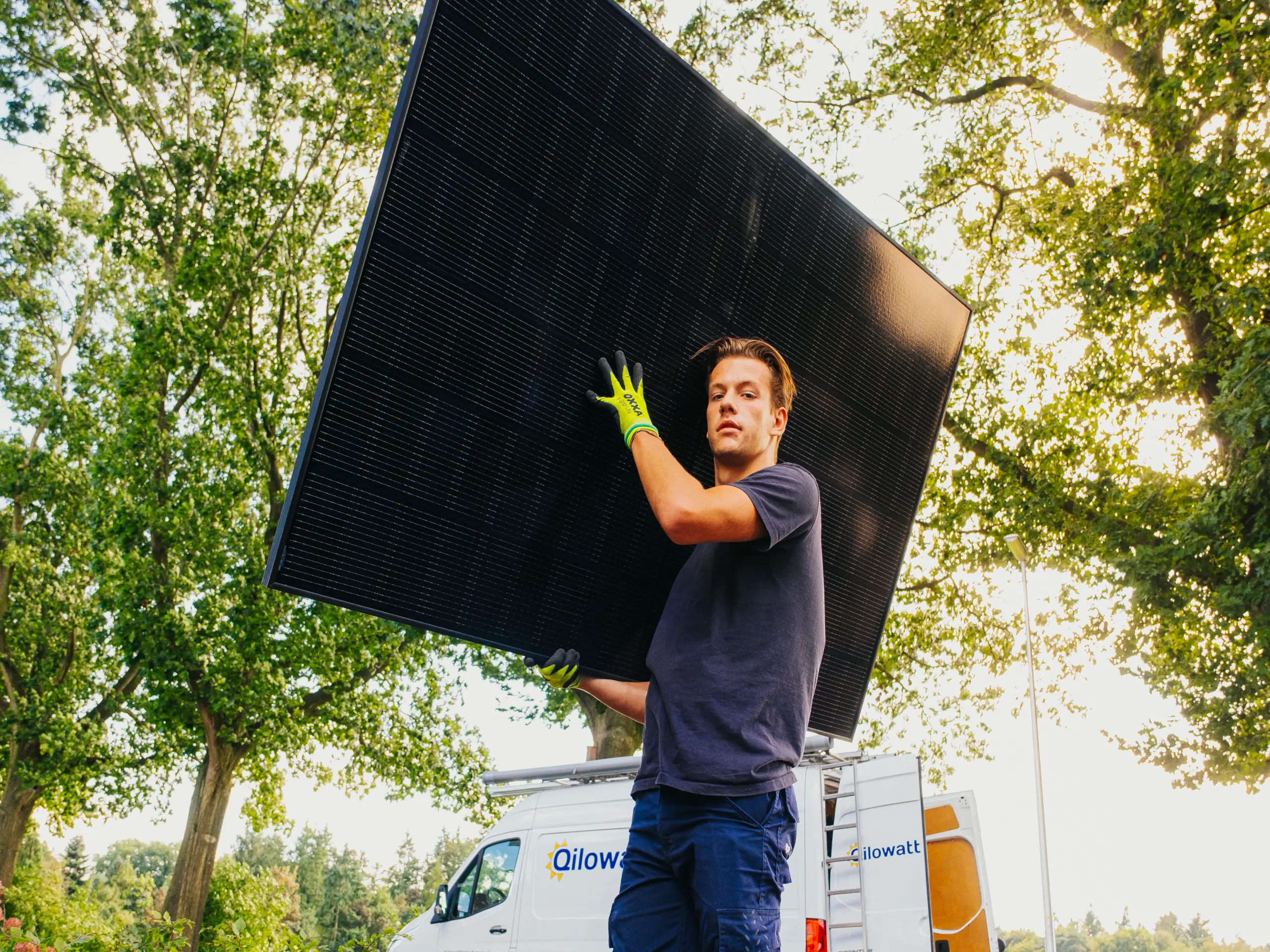 Man draagt een groot zonnepaneel buiten, met een Qilowatt bestelwagen op de achtergrond.