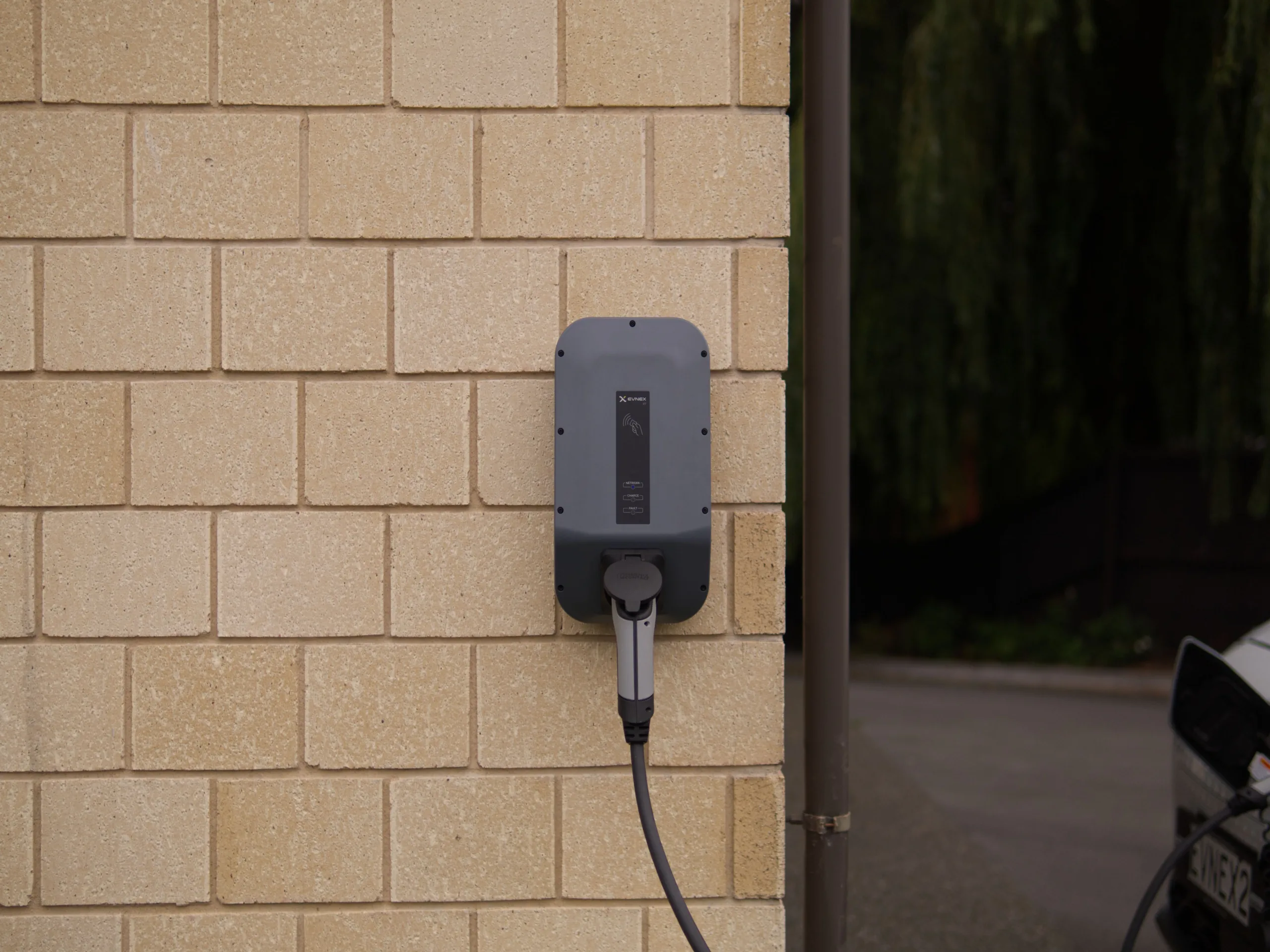 Elektrische laadpaal bevestigd aan een beige bakstenen muur met een kabel aangesloten.