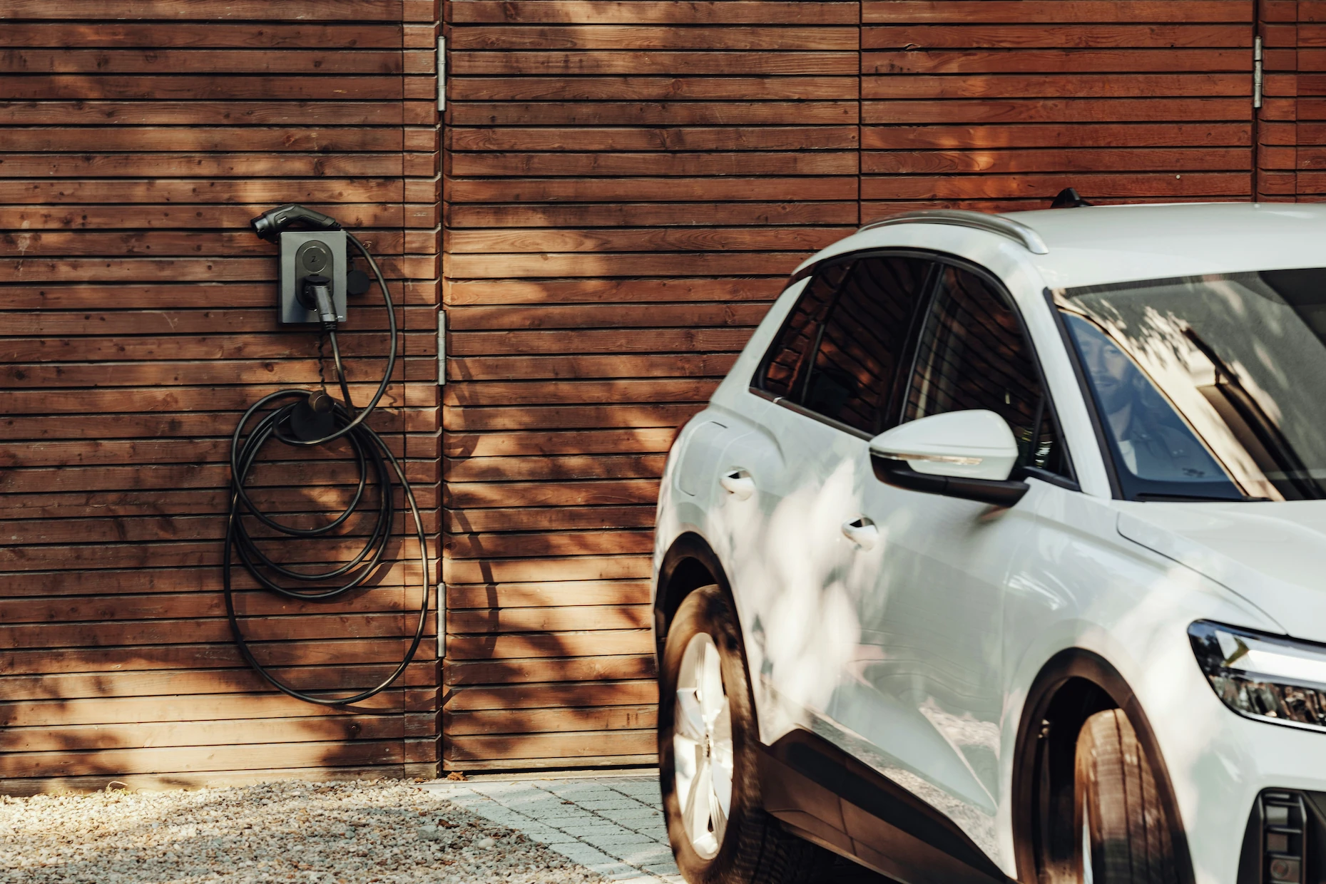 Witte elektrische auto geparkeerd naast een houten muur met een wandlader en kabel.