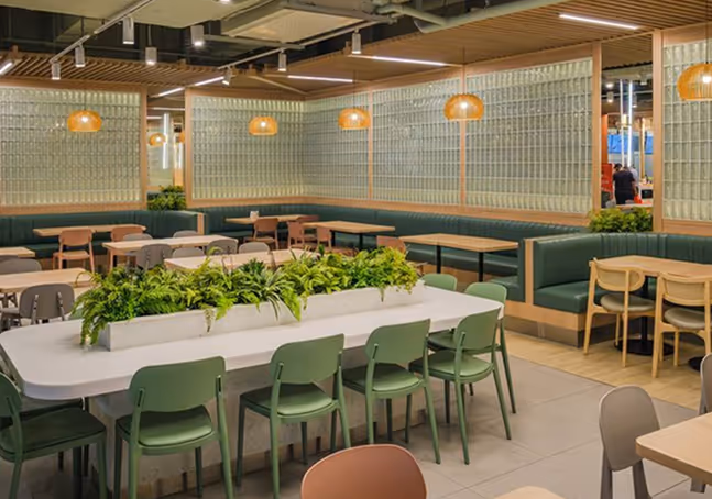 Modern cafeteria with green chairs, wooden tables, booth seating along glass block walls, and hanging pendant lights.