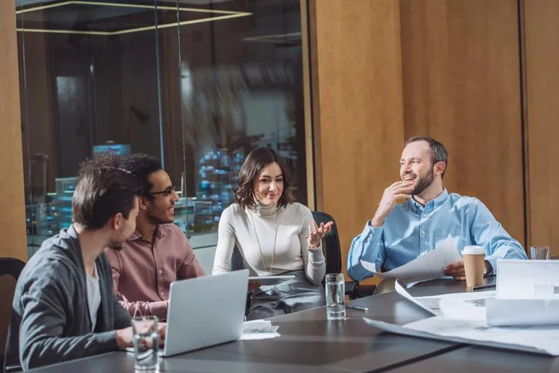 Groupe de collègues en réunion autour d’une table, discutant et souriant dans une salle de réunion moderne.