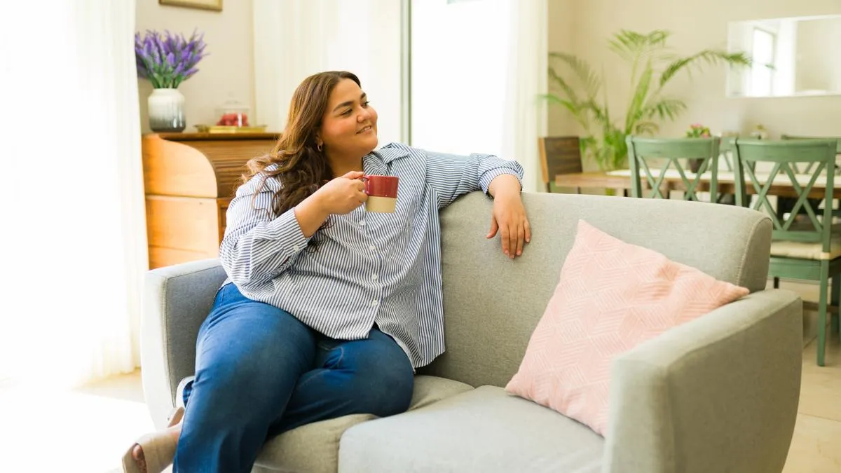 Femme assise confortablement sur un canapé dans un salon lumineux, tenant une tasse et profitant d’un moment de détente.