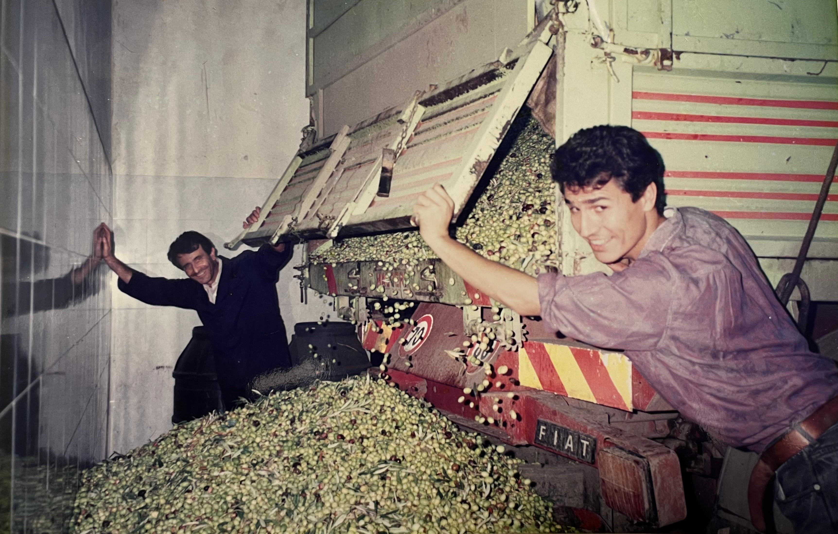 Interno di oleificio storico con macchinari industriali in metallo blu e due uomini che discutono vicino alle macchine.